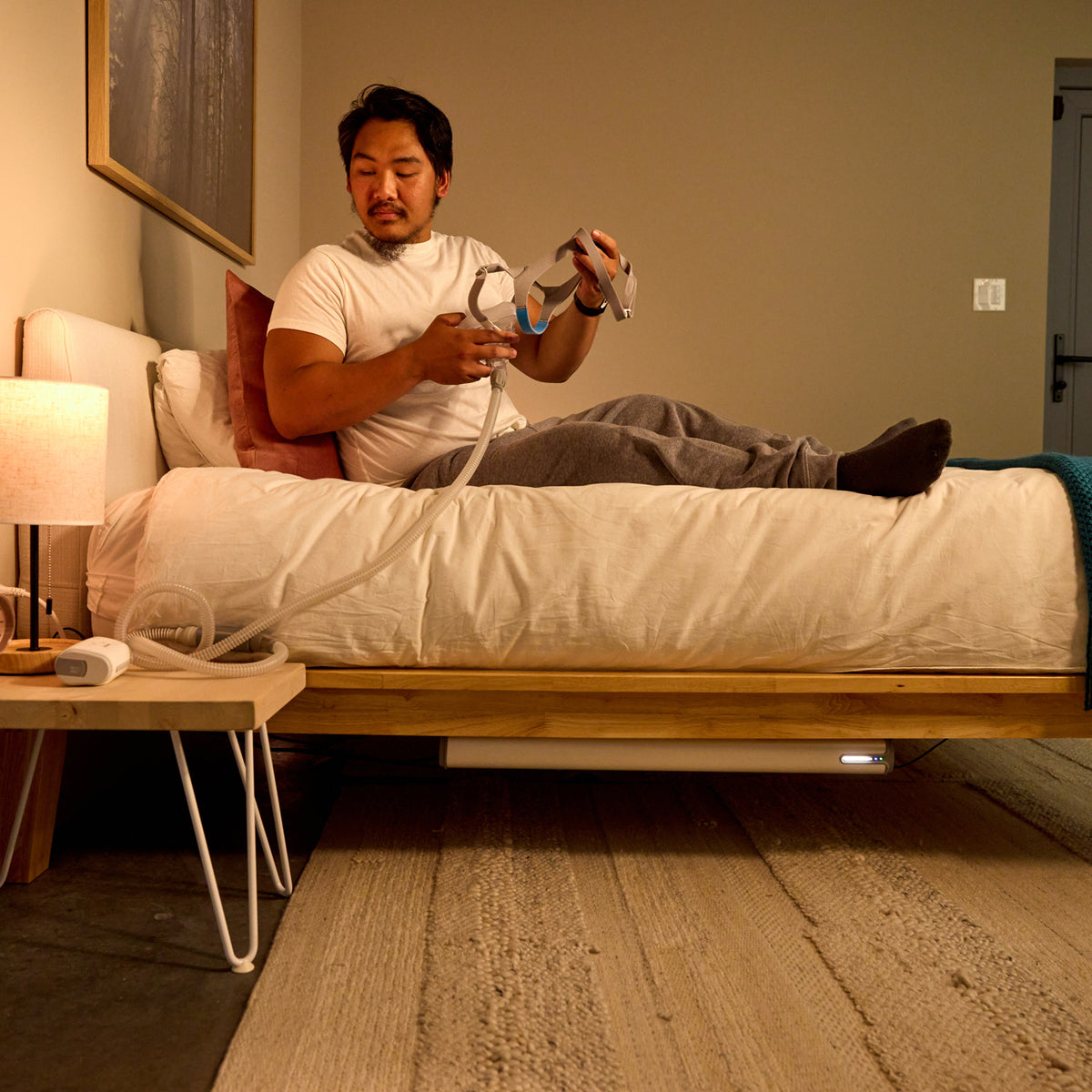 A man lies on a bed in a bedroom while using a white and gray CPAP. The room has beige walls, a wooden bed frame, a bedside table with a lit lamp, and a textured rug on the floor. Under the bed there is a Backup Core power station powering the CPAP device.