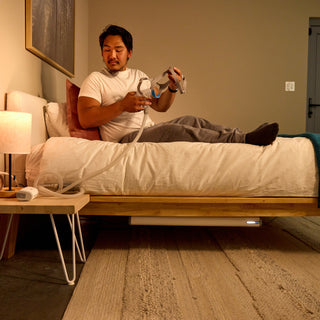 A man lies on a bed in a bedroom while using a white and gray CPAP. The room has beige walls, a wooden bed frame, a bedside table with a lit lamp, and a textured rug on the floor. Under the bed there is a Backup Core power station powering the CPAP device.
