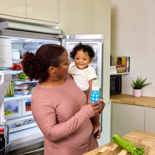A woman in a dusty rose long-sleeve shirt holds a smiling toddler wearing a white outfit while standing in a kitchen next to an open refrigerator with the Backup Core battery backup system installed on top of the fridge. The toddler is holding a blue and white polka-dot bottle.