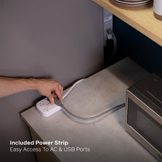 A hand plugging a white power strip into a countertop outlet near a microwave, with text reading 'Included Power Strip' and 'Easy Access To AC & USB Ports'. The power strip is connected to the Backup Core Power Station and is powering the fridge and the microwave.
