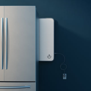 A sleek wall-mounted power station (Backup Core) with the BioLite logo, shown plugged into an outlet next to a stainless steel refrigerator against a dark blue wall.